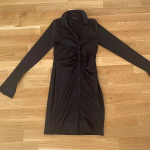 Svart skjortklänning från NLY ONE XS -  Svart skjortklänning från NLY ONE i storlek XS. Klänningen har långärmade ärmar, krage, knappar hela vägen framtill och snygg rynkdetalj vid midjan. Materialet är mjukt och stretchigt, vilket gör den bekväm och smickrande. Perfekt för dig som gillar stilrena och trendiga plagg.