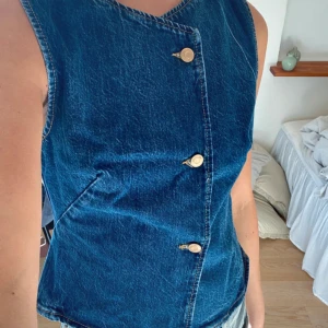 Blå jeansväst från Zara med knappar - Snygg blå jeansväst från Zara med tre guldfärgade knappar framtill och rund halsringning. Västen har en asymmetrisk knäppning och två diskreta fickor på sidorna. Perfekt att styla över en t-shirt eller skjorta för en cool look.