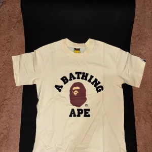 A Bathing Ape beige t-shirt - Beige t-shirt från A Bathing Ape med klassisk ape-logga i brunt och svart tryck på bröstet. Rund hals och korta ärmar, samt en liten ape-logga på ärmslutet. Tillverkad i mjuk bomull för skön passform och streetwear-vibe.
