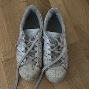 Adidas Superstar vita sneakers - Klassiska Adidas Superstar sneakers i vitt med grå snörning och ikoniska tre ränder på sidorna. Skorna har rund tå och platt sula med den typiska snäckformade tåkappan. Materialet är skinn och textil på insidan. Perfekt för dig som gillar retrostil och streetwear.