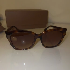 Burberry solglasögon brun mönstrad - Snygga solglasögon från Burberry med brunmelerat mönster och bred båge i cat eye-form. Guldigt Burberry-logo på sidan och mörka glas. Kommer med originalfodral och box. Perfekt accessoar för att lyfta din stil.