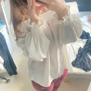 Vit offshoulder blus med volang - Supersöt vit blus med offshoulder och bred smock upptill. Långa ärmar med smock och volang vid ärmslut. Säljer då den inte kommer till användning längre.