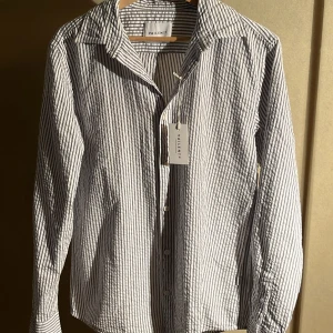 Spiller ny Randig skjorta från Vailent - Snygg skjorta från Vailent med smala vertikala ränder i vitt och grått. Klassisk krage, knäppning framtill och långa ärmar. Tillverkad i bomull, perfekt för en clean och stilren look.