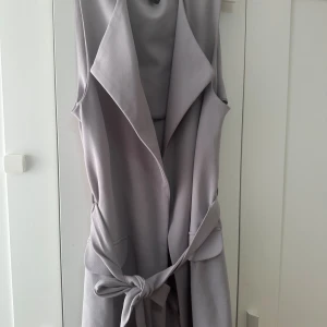 Ljusgrå lång väst med knytband - Snygg ljusgrå väst med breda slag och knytband i midjan. Västen är ärmlös och har fickor framtill, perfekt för att lyfta vilken outfit som helst. Stilren och modern design med en längre passform.