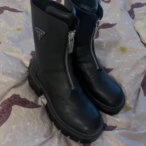 Svarta boots från Guess - Chunky svarta boots från Guess i skinn med grov sula och silverfärgad dragkedja framtill. Klassisk rund tå och Guess-logga i metall på sidan. Perfekta för dig som vill ha en edgy och stilren look. Storlek 38/39 aldrig används bara testade m, nypris va runt 3000 kr
