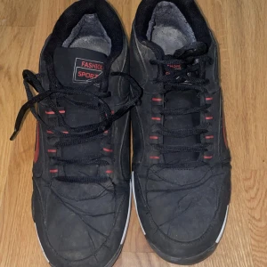 Svarta sneakers med röda detaljer - Svarta sneakers med röda detaljer och snörning. Skorna har en sportig look med rund tå, platt sula och röda accenter längs sidorna och snörningen. Ovandelen är i syntetmaterial och insidan är fodrad för extra komfort. Perfekta för dig som gillar en klassisk streetstil.