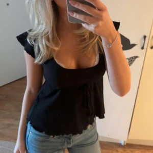 Svart blus med volangärmar och knyt - Säljer en svart blus med djup v-ringning och volangärmar. Toppen har knyt under bysten och en lätt croppad passform. Detaljerad med vit spetskant vid halsen som ger en extra touch. Perfekt att styla med jeans för en cool look.