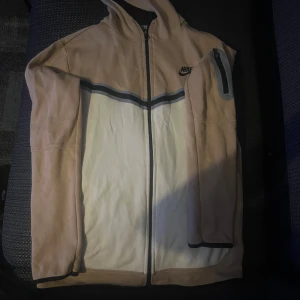 Nike beige och vit vindjacka med huva - Nike hoodie i beige och vitt med svart logga på bröstet. hoodie har hel dragkedja, huva och en cool ficka med dragkedja på ärmen. Snygga svarta och grå detaljer över bröstet. Perfekt för chill och varmt.