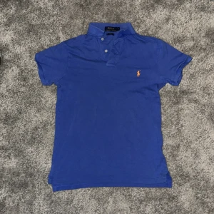 Blå Polo Ralph Lauren pikétröja S - Blå pikétröja från Polo Ralph Lauren i slim fit med klassisk krage och två knappar framtill. Tröjan har det ikoniska orange broderade Polo-logot på bröstet. Tillverkad i mjuk bomull som är skön mot huden och passar perfekt till jeans eller shorts.