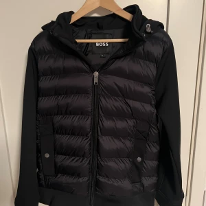 Svart cardigan från BOSS med huva - Svart cardigan från BOSS i storlek S. Jackan har en snygg quiltad framsida, huva med snörning och dragkedja framtill. Ärmarna är släta och materialet är en mix av polyester och syntet. Perfekt för dig som gillar stilren och sportig look. Använd några gånger, inga defekter eller sådant! Priset kan diskuteras och är ej hugget i sten! 