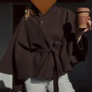 Brun kappa med bälte och vida ärmar - Säljer en brun kappa med högt krage, vida ärmar och knytbälte i midjan. Jackan har en stilren look med knappar framtill och är perfekt för dig som gillar minimalistisk design och vill ha något som sticker ut. Passar dig som gillar att klä dig trendigt och bekvämt. Knappt använd💞