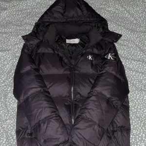 Svart pufferjacka från Calvin Klein - Svart pufferjacka från Calvin Klein Jeans med huva och dragkedja. Jackan har CK-logga på bröstet och ett stort CK-märke på ärmen. Fylld med syntetmaterial och har en klassisk, quiltad design. Perfekt för kalla dagar och har en normal passform.