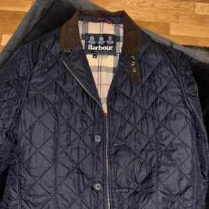 Mörkblå quiltad jacka från Barbour - Snygg mörkblå quiltad jacka från Barbour med klassiskt rutigt foder och bruna detaljer på kragen. Jackan har både dragkedja och knappar framtill, samt röda kantband på insidan. Perfekt för dig som gillar stilren och tidlös design.