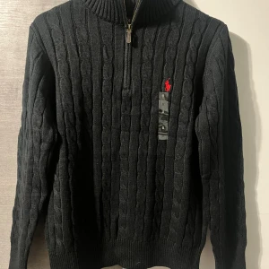 Polo Ralph Lauren half zip - Orginal pris: 2113kr, endast för 899kr hos Luxe Vibes! Svart kabelstickad tröja från Polo Ralph Lauren med halv dragkedja och röd broderad logga på bröstet. Tröjan har hög krage, ribbade muddar och är tillverkad i mjukt stickat material. Perfekt för lager-på-lager under höst och vinter.