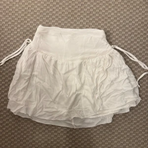 Vit volangkjol med snörning - Superfin vit kortkjol med volanger och snörning i sidorna. Kjolen har en hög midja och dragkedja bak för enkel påklädning. Tunt, luftigt tyg som passar perfekt till varma dagar. Stilren och enkel att matcha med olika toppar.