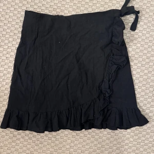 Svart omlottkjol med volang - Svart omlottkjol med knytband i midjan och volang längs nederkanten. Kjolen har en enkel och stilren design som passar perfekt till många olika looks. Tillverkad i ett mjukt tyg som känns skönt mot huden.