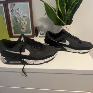 Nike Air Max 90 svart/vit strl 45 - Säljer ett par klassiska Nike Air Max 90 i svart och vitt. Skorna har en chunky sula med synlig air-bubbla, mesh och skinnpaneler samt vita swoosh-loggor på sidorna. Perfekta för dig som gillar streetwear och vill ha en ikonisk sneaker i garderoben.