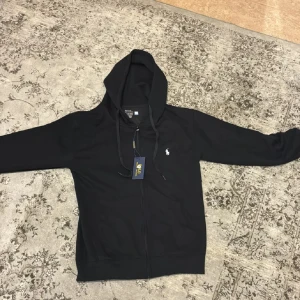 Svart hoodie från Polo Ralph Lauren  - Svart hoodie från Polo Ralph Lauren med klassisk vit logga på bröstet. Tröjan har huva med snörning, dragkedja framtill och långa ärmar. Tillverkad i mjuk bomull för skön känsla. Perfekt för chill dagar och streetwear-stil. Priset är för hela settet 