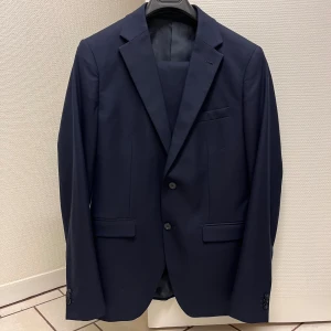Mörkblå kostym från Brothers - Säljer en stilren mörkblå kostym från Brothers, modell Jonah Stretch i slim fit, av märket Riley. Kavajen har två knappar, klassiska slag och fickor med lock. Byxorna är raka och matchar kavajen perfekt. Perfekt för dig som vill ha en clean och modern look. Storlek 50. Inköpt på Brothers 2023. Använd vid två tillfällen. Kemtvättad