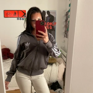 Grå Adidas vindjacka med ränder - Säljer en grå Adidas vindjacka med klassiska blåa ränder längs ärmarna och Adidas-logga på bröstet. Jackan har dragkedja framtill, ribbade muddar och krage. Perfekt för dig som gillar sportig och chill stil.