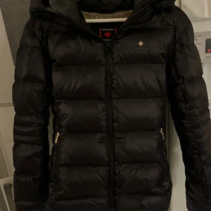 Svart pufferjacka från Canada Snow - Säljer en svart pufferjacka från Canada Snow med huva och guldfärgad logga på bröstet. Jackan har dragkedja framtill och två sidofickor med dragkedja. Insidan är beige med tryckt logga. Perfekt för kalla vinterdagar.