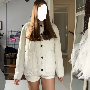 Vit quiltad pufferjacka från Zara - Quiltad pufferjacka från Zara i ljusvit färg med svarta knappar framtill. Jackan har två stora fickor, rund hals och långärmad passform. Perfekt för dig som gillar en clean och chill stil. Materialet är mjukt och känns skönt mot huden.