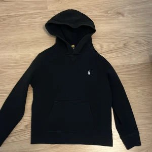 Svart hoodie från Polo Ralph Lauren - Svart hoodie från Polo Ralph Lauren med klassisk huva och magficka. Tröjan har en liten vit broderad logga på bröstet och är tillverkad i mjukt material. Perfekt för en chill och stilren look. Denna köptes för 1195 och har använts fåtal gånger därför säljer jag denna för 449+frakt hör av er vid frågor och pris!😊