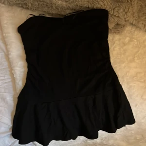 Svart tubtopp med volang  BikBok - Supersnygg svart tubtopp med volang från BikBok med axelbandslös design och volang nertill. Tillverkad i ett mjukt och lätt material från EcoVero. Perfekt för dig som vill ha en enkel men trendig look.