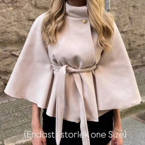 Beige capejacka med bälte och knapp - Snygg beige capejacka med hög krage, dekorativ guldig knapp vid halsen och knytskärp i midjan. Jackan har vida ärmar och en elegant, flowy siluett som ger en cool och trendig look.