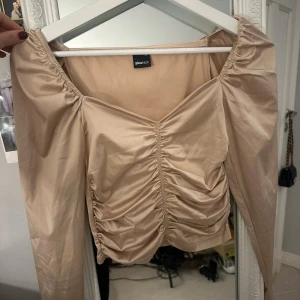 Beige omlottopp från Gina Tricot - Snygg beige omlottopp från Gina Tricot i glansigt material. Toppen har långa ärmar, v-ringning och rynkade detaljer framtill som ger en cool look. Perfekt för dig som gillar trendiga och stilrena plagg med det lilla extra.