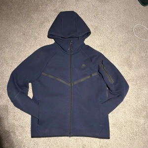 Mörkblå Nike Tech Fleece dragkedja - Nike Tech Fleece hoodie i mörkblått med hel dragkedja, huva och diskret Nike-logga på bröstet. Jackan har en snygg diagonal detalj över bröstet och en praktisk ficka med dragkedja på ärmen. Perfekt för en sportig och clean look.