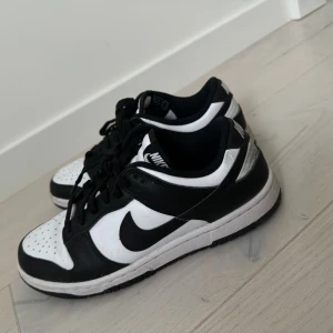 Nike Dunk Low svartvita sneakers - Nike Dunk Low sneakers i svart och vitt med klassisk swoosh på sidorna. Skorna har snörning, perforerad tå och låg profil. Tillverkade i skinn och syntetmaterial, perfekt för dig som gillar streetstyle och vill ha en ikonisk look.
