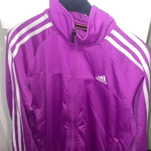 Lila Adidas vindjacka med dragkedja - Snygg lila vindjacka från Adidas med klassiska vita ränder längs ärmarna och Adidas-logga på bröstet. Jackan har hel dragkedja framtill och hög krage. Perfekt för dig som gillar sportig stil och vill sticka ut.
