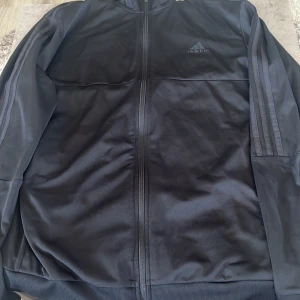 Svart Adidas vindjacka med dragkedja - Svart vindjacka från Adidas med klassiska tre ränder längs ärmarna och broderad logga på bröstet. Jackan har hel dragkedja framtill, ribbade muddar och är tillverkad i lätt polyester. Perfekt för dig som gillar sportig stil och streetwear. Nästan aldrig använda! 10/10 skick.