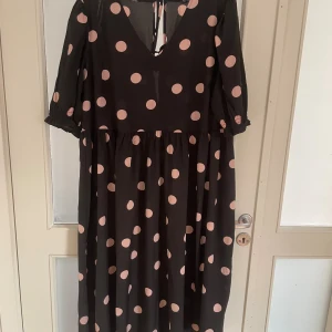 Svart prickig långklänning New Look - Svart långklänning från New Look Curves med stora ljusrosa prickar. Klänningen har v-ringning, knytning i ryggen och halvlånga puffärmar med resår. Tillverkad i ett lätt och luftigt syntetmaterial som ger ett flowigt fall.