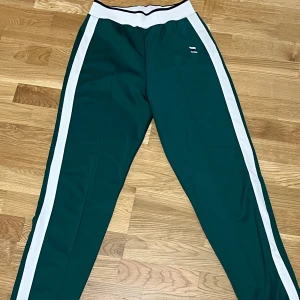 Gröna träningsbyxor - Snygga gröna träningsbyxor från Zara med vita sidoremsor och bred vit midja med svart detalj. Byxorna har en sportig look och är gjorda i ett mjukt syntetmaterial som är perfekt för chill eller träning.