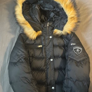 Svart dunjacka Rockandblue med fuskpäls - Snygg svart dunjacka från Rockandblue med quiltade detaljer och avtagbar huva med fuskpäls. Jackan har flera dragkedjefickor, Rockandblue-logga på ärmen och stängs med både dragkedja och knappar. Perfekt för kalla dagar och riktigt street.