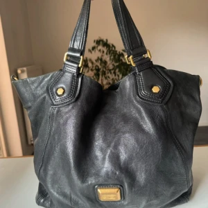 Svart skinnväska från Marc by Marc Jacobs - Cool svart handväska i äkta lammnappa från Marc by Marc Jacobs. Väskan har guldfärgade metalldetaljer och ett unikt innerfoder med rött och svart stjärnmönster. Rymlig modell med dubbla handtag, finns visst slitage men inte mycket märkbart.