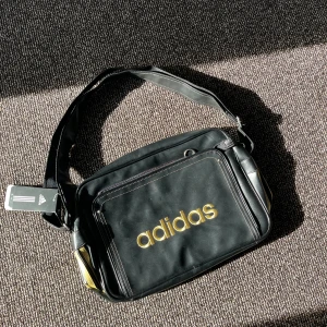 Adidas väska - Snygg retroinspirerad axelväska från Adidas med guldigt tryck framtill. Väskan har justerbar axelrem, stort huvudfack med dragkedja och flera mindre fack för bra förvaring. Perfekt till vardag, skola eller gym. I nytt skick, aldrig använd Rymlig modell – plats för laptop, böcker m.m. Material: Slitstark syntet (läderimitation)