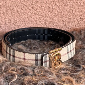 Rutigt skinnbälte med guldspänne - Säljer ett snyggt Burberry bälte i skinn med klassiskt rutigt mönster i beige, svart och röd. Bältet har ett stilrent guldspänne med Burberrys ikoniska B-logga. Insidan är svart med präglat Burberry-motiv. Perfekt för att lyfta din outfit. Tror längd 105cm. 