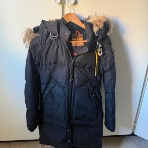 Svart Parajumpers lång dunjacka med päls - Säljer en svart lång dunjacka från Parajumpers med ikonisk gul rem och patch på ärmen. Jackan har huva med fluffig päls, dragkedja och stora fickor framtill. Perfekt för kalla dagar och riktigt snygg streetstil. Storlek XS men passar S också 