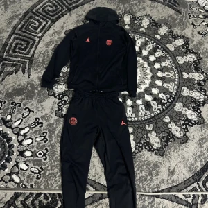 Jordan PSG svart träningsset med huva - Svart träningsset från Jordan och PSG med röda loggor på både jacka och byxor. Jackan har huva, hel dragkedja och fickor. Byxorna har resår i midjan och smal passform. Perfekt för dig som gillar sportig stil och vill representera PSG.