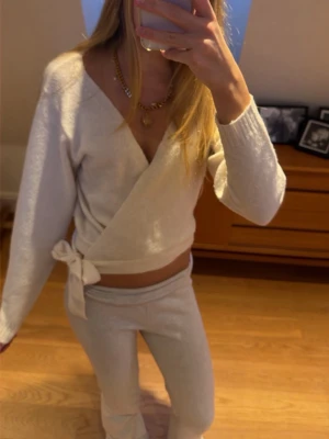 Vit omlottkofta med knyt i midjan - Mysig vit omlottkofta med knyt i midjan och djup v-ringning. Tröjan är långärmad och har en croppad passform som ger en avslappnad men ändå trendig look. Perfekt att styla med låga byxor eller kjol för en chill vibe.