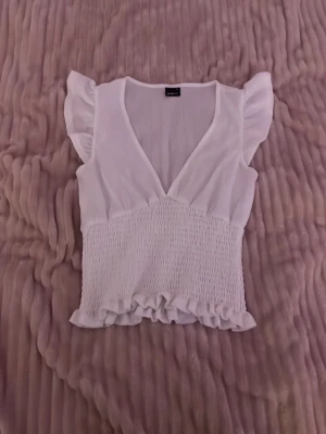 Vit blus med volang och v-ringning - Säljer en söt vit blus från Ginatricot med v-ringad hals, volangdetaljer vid axlar och nederkant samt smockad midja. Blusen är ärmlös och har en luftig, feminin look som passar perfekt till sommaren.