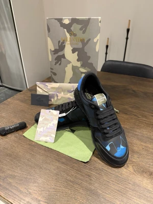 Valentino Garavani Rockrunner sneakers - Säljer ett par Valentino Garavani Rockrunner sneakers i svart och blått med coolt kamouflagemönster. Skorna har snörning, rund tå och platt sula. Materialmix av skinn och textil ger en lyxig känsla. Kommer med originalkartong, extra skosnören och dustbag.