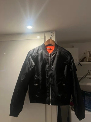 Svart häst bomberjacka från Copperfield - Säljer en svart rid bomberjacka från Copperfield med orange innerfoder och broderad detalj på ärmen. Jackan har dragkedja framtill, fickor med knapp och ribbade muddar vid krage och ärmslut. Storlek 170cm