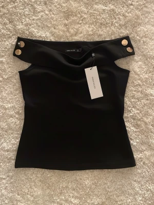 Svart axelbandslös topp Karen Millen - Snygg svart axelbandslös topp från Karen Millen med bred offshoulder-krage och dekorativa guldknappar på axlarna. Toppen har en figurnära passform och är tillverkad i ett slätt, stretchigt material som ger en stilren look.