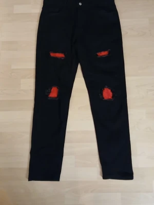 Svarta jeans från Temu med röda slitningar XL - Svarta jeansbyxor i storlek XL med coola röda detaljer bakom slitningarna på knäna och låren. Byxorna är raka i modellen och tillverkade i en mix av bomull, polyester och elastan för skön stretch. Perfekta för dig som vill sticka ut lite extra. Från Temu!