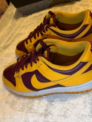 Nike Dunk Low gul och vinröd - Nike Dunk Low sneakers i en riktigt snygg färgkombo av gul och vinröd. Skorna har klassisk låg siluett, vit sula och matchande vinröda skosnören. Tillverkade i skinn med tydliga Nike-loggor på häl och tunga. Svårt och hitta med bra pris, men perfekt för dig som vill sticka ut med färg!