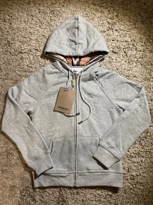 Snygg ljusgrå hoodie från Burberry - Snygg ljusgrå hoodie från Burberry med klassiskt rutigt foder i huvan, broderad logga på bröstet och hel dragkedja. Tröjan har snörning i huvan och praktiska fickor framtill. Perfekt för dig som vill ha en stilren och exklusiv look.